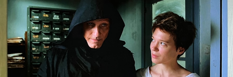A man in a black hood with a woman in a white dress half-smiling at him.