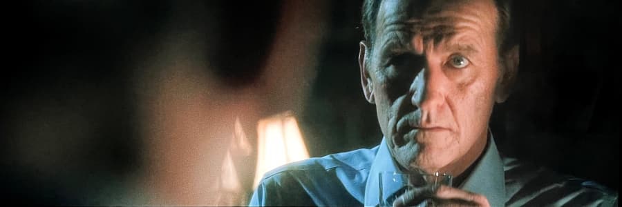 An older man looks at himself drinking a glass of whiskey in the mirror