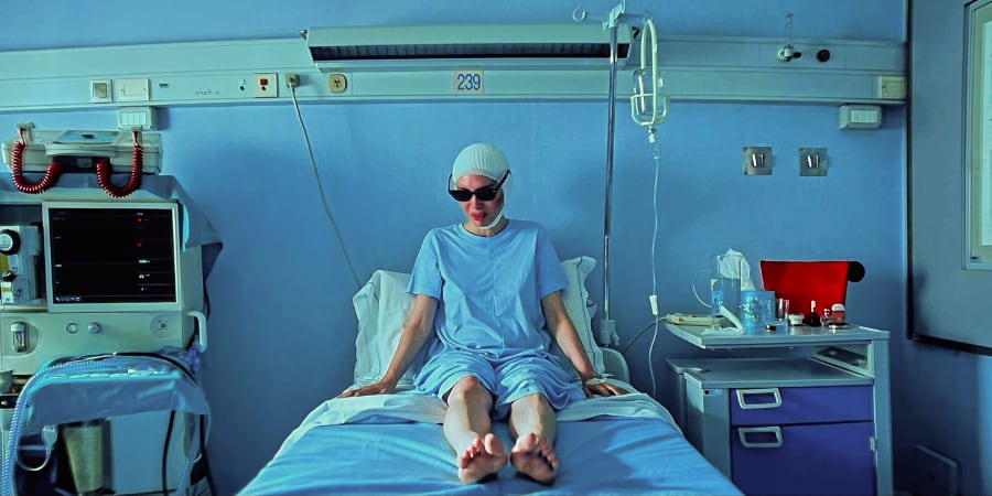 A woman sits in a hospital bed with bandages around her head and wearing dark glasses.