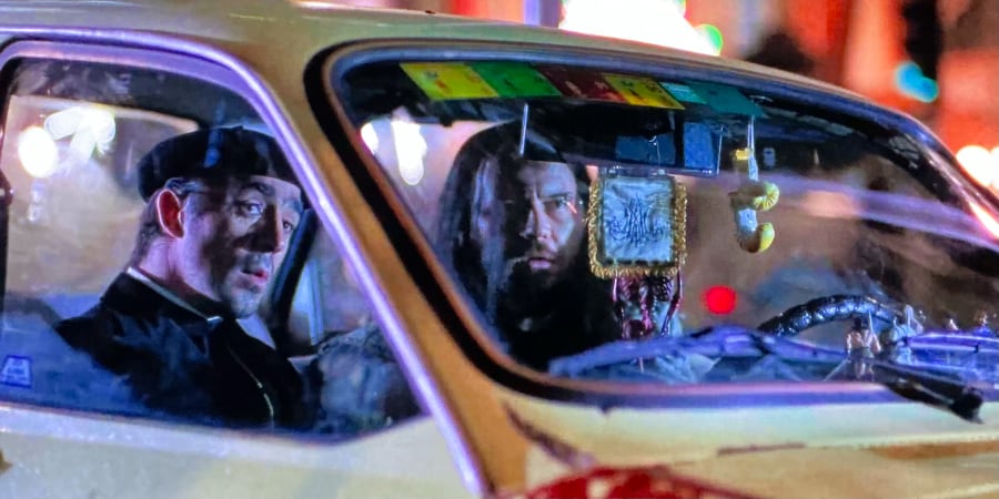 A priest and a metalhead sit in a car watching for something.