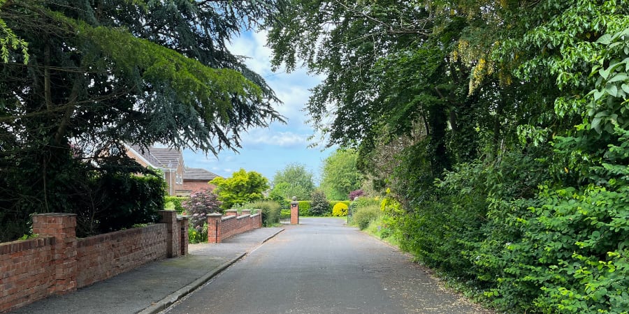 A road leading to gates to with trees on the right and houses on the left.