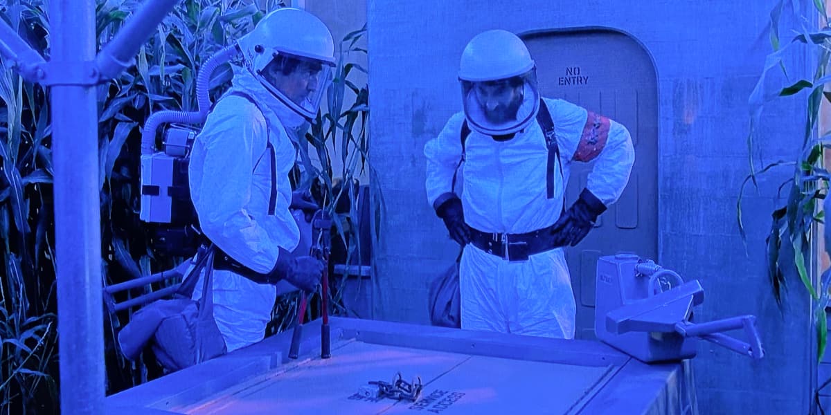 Two men in hazard suits in a greenhouse with blue lighting.