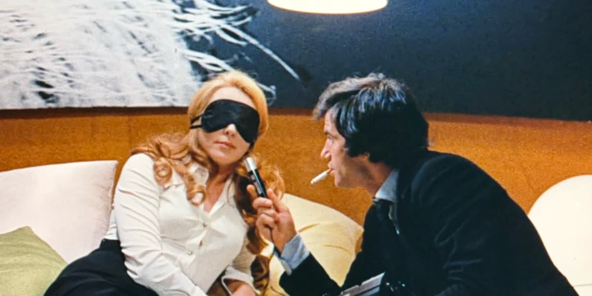 A man holds a microphone to the mouth of a beautiful woman wearing a blindfold.
