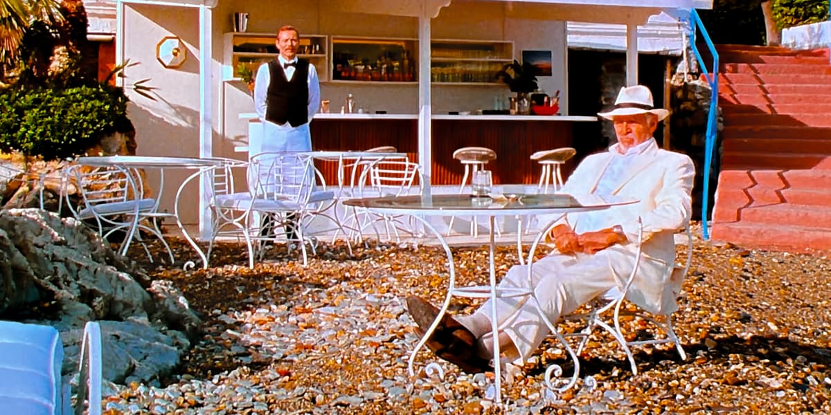 An old man in a white suit sips a drink on a beach and looks pensively out at sea.