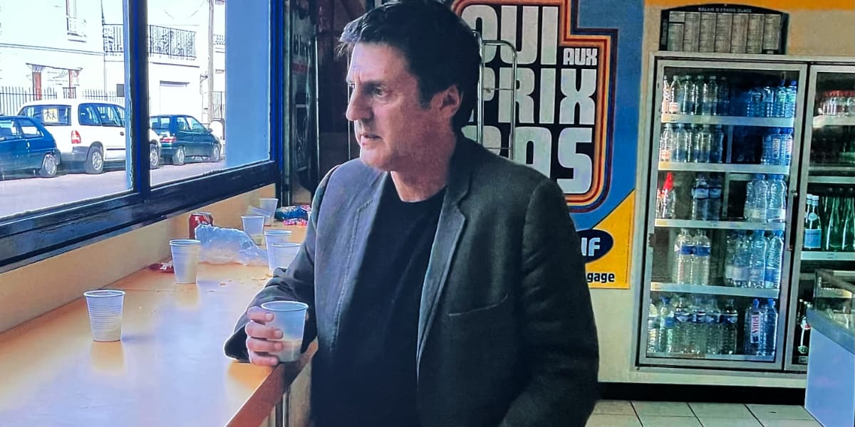 A man looks pensively out of a shop window holding a plastic cup of coffee.