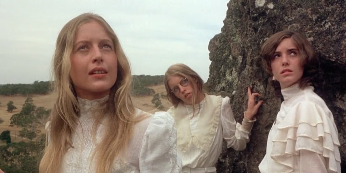 Three girls in white dresses look up at Hanging Rock,