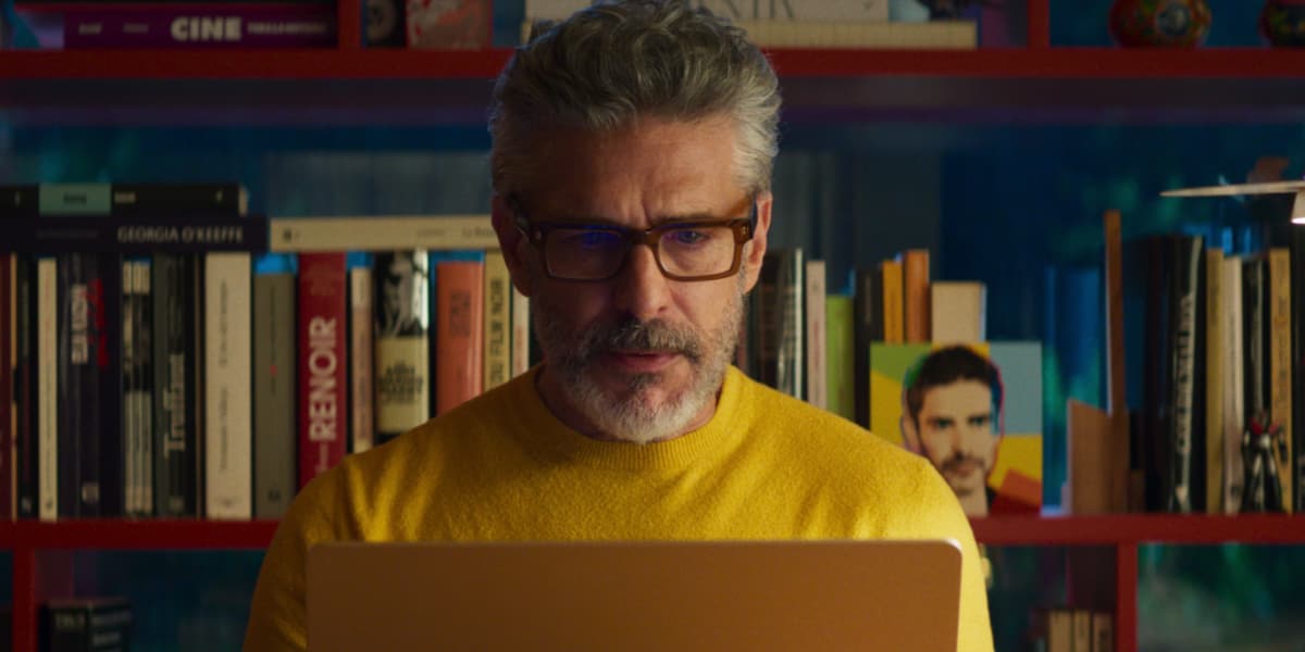 A man sits at his desk typing on his laptop wearing a yellow jumper.