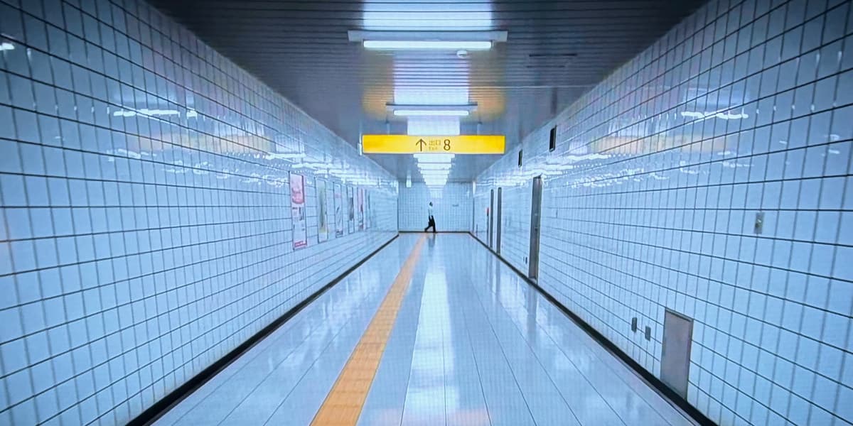 A man smiles in a deranged way at the camera in a white-tiled Tokyo subway corridor.