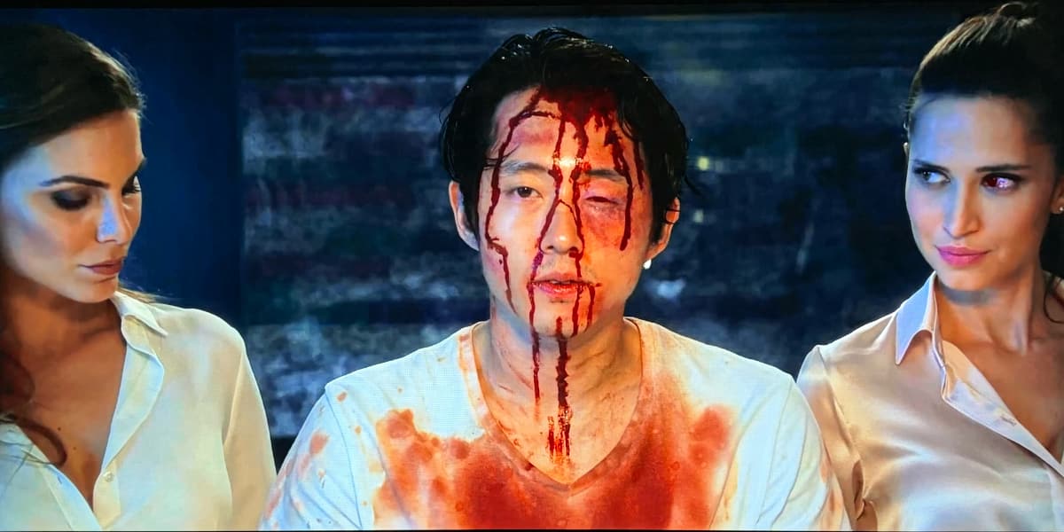 A man, bleeding from the head, is flanked by two pristine, beautiful women in business suits.